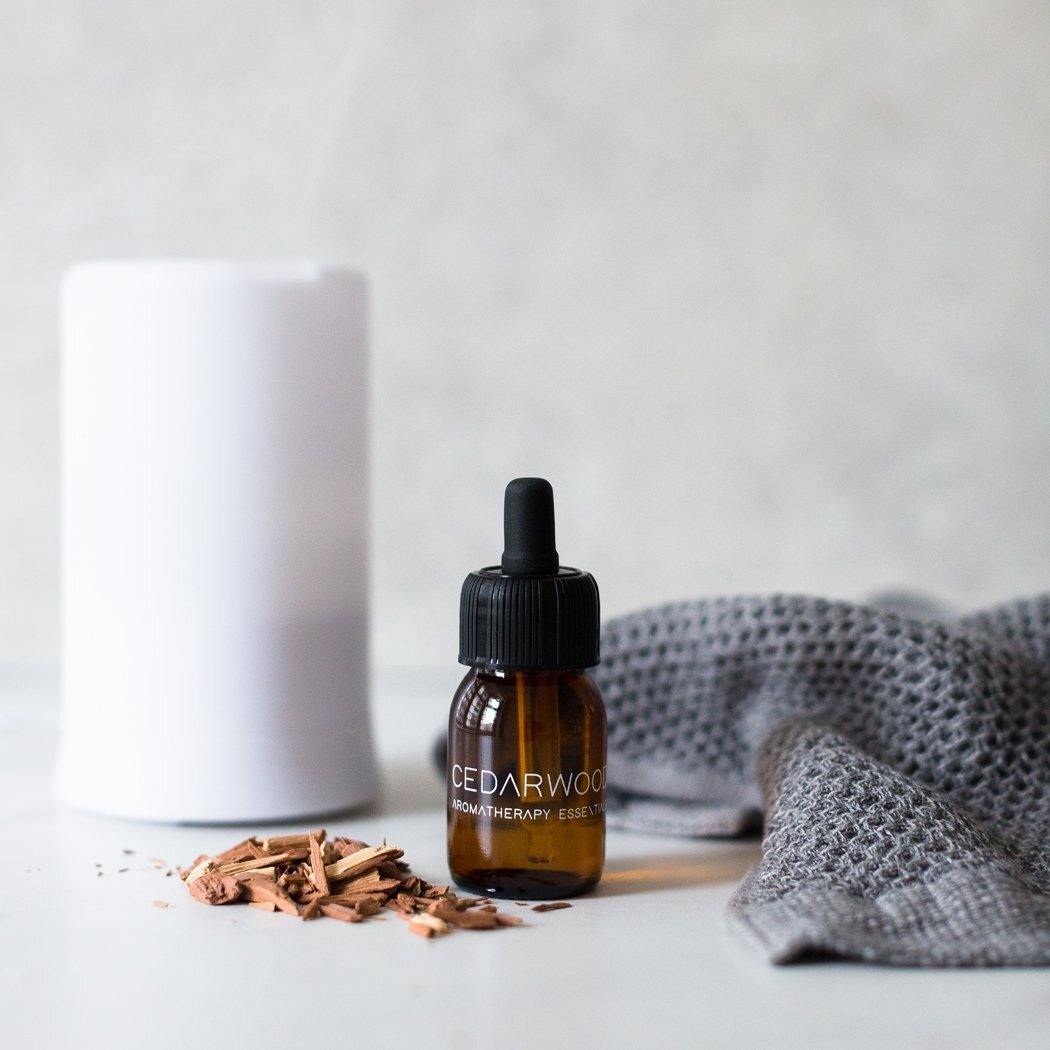 Een flesje RainPharma Essential Oil Cedarwood rust naast houtkrullen, een grijze doek met textuur en een witte geurverspreider op een licht oppervlak, wat de kalmerende cedergeur oproept.