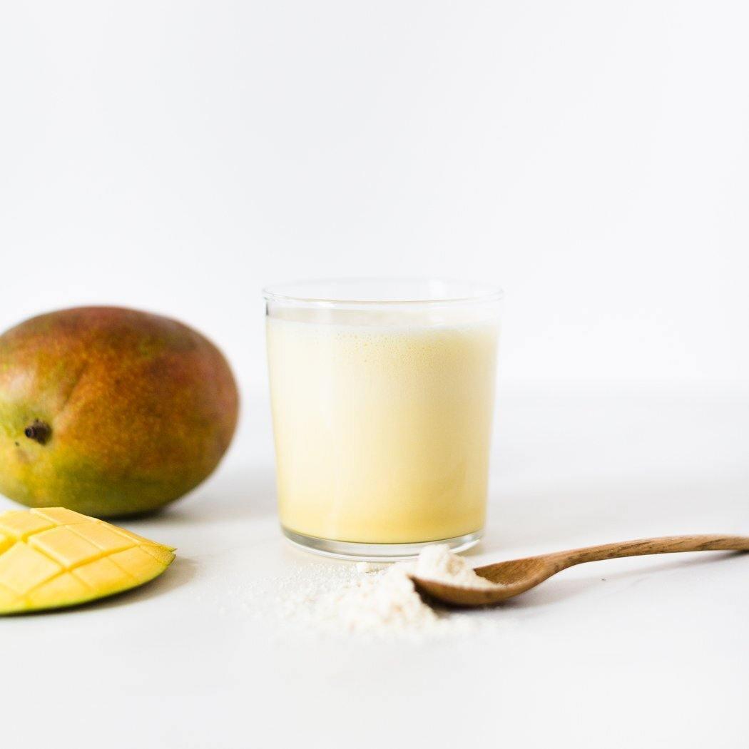 Een glas Exotic Mango-go Shake met een gesneden mango en een hele mango ernaast, en een houten lepel met Rainpharma mangopoeder op een witte achtergrond, alles voor een vegetarisch genot.