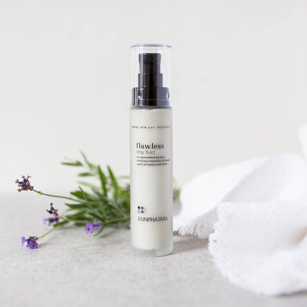 Een flesje RainPharma Flawless Day Fluid - Dagcrème onzuivere van vette huid staat op een licht oppervlak, samen met een takje lavendel, wat groen en een witte doek.