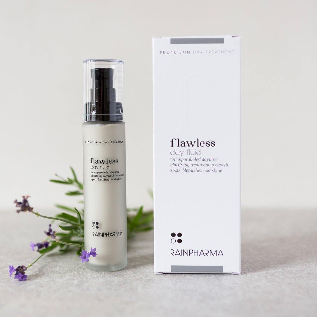 Een flesje en doosje RainPharma Flawless Day Fluid - Dagcrème onzuivere van vette huid zijn afgebeeld op een licht oppervlak met takjes lavendel op de achtergrond.