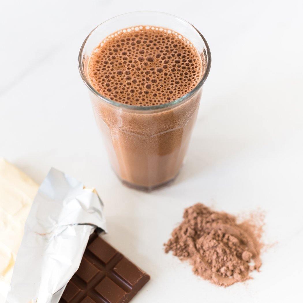 Een glas verleidelijke RainPharma Shake Melkchocolade staat naast een gedeeltelijk uitgepakte chocoladereep en een berg rijke cacaopoeder op een onberispelijk wit oppervlak.