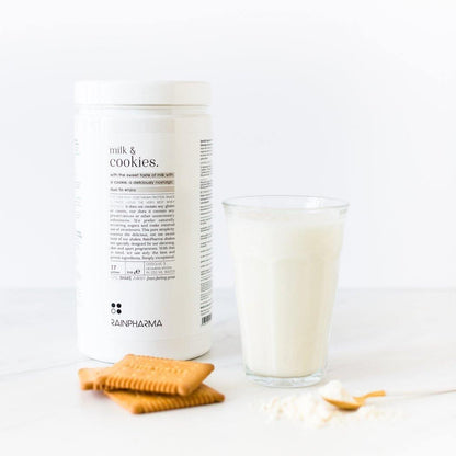 Een RainPharma Shake Milk&amp;Cookies pot staat naast een glas melk, twee biscuits en een klein houten lepeltje met poeder op een wit oppervlak en biedt een eiwitrijk tussendoortje dat perfect is voor elk moment van de dag.