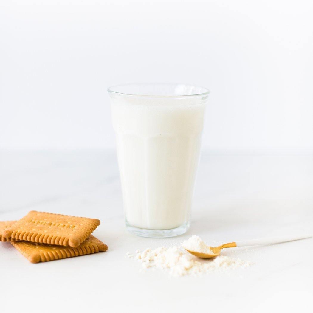 Een glas RainPharma Shake Milk&amp;Cookies staat op tafel naast twee vierkante koekjes en een lepeltje poeder, waardoor een eiwitrijk tussendoortje ontstaat.