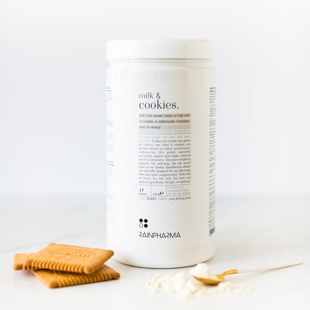 Een verpakking RainPharma Shake Milk&amp;Cookies van Rainpharma staat naast vier vierkante koekjes en een klein houten bolletje gevuld met proteïnepoeder op een maagdelijk wit oppervlak.