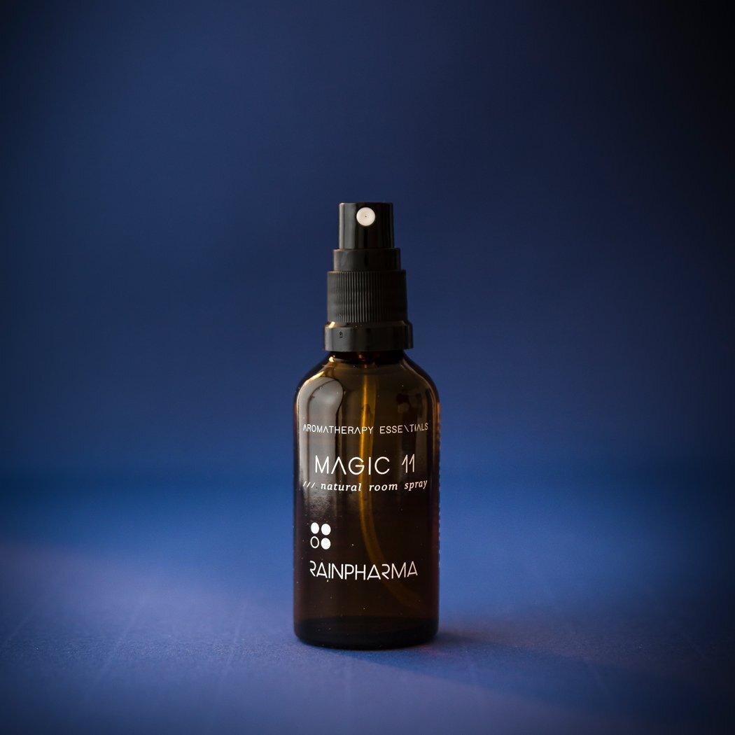 Een bruine glazen fles met het label "Natural Room Spray - Magic 11" van RainPharma, gemaakt met essentiële oliën, staat tegen een donkerblauwe achtergrond.