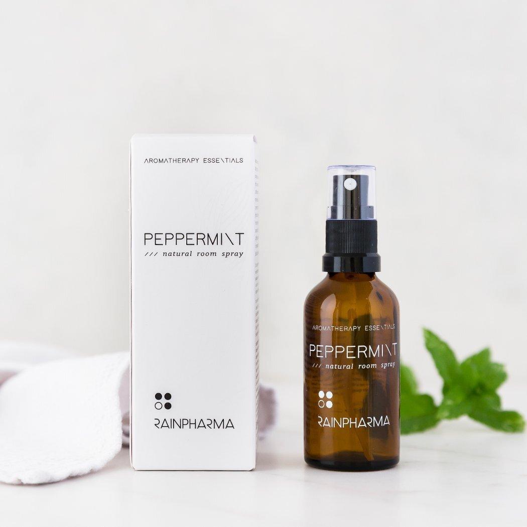 Een bruine glazen RainPharma Natural Room Spray Peppermint fles staat naast zijn witte doos, die beide het gebruik van essentiële oliën voor een natuurlijke huisgeur benadrukken. Op de achtergrond is een muntblaadje zichtbaar.