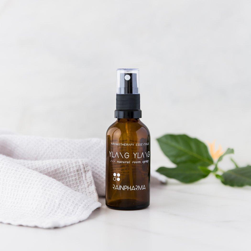 Een bruin glas RainPharma Natural Room Spray - Ylang Ylang rust op een wit oppervlak naast een opgevouwen handdoek en een groene bladplant - perfect als een natuurlijk huisparfum met essentiële olie.