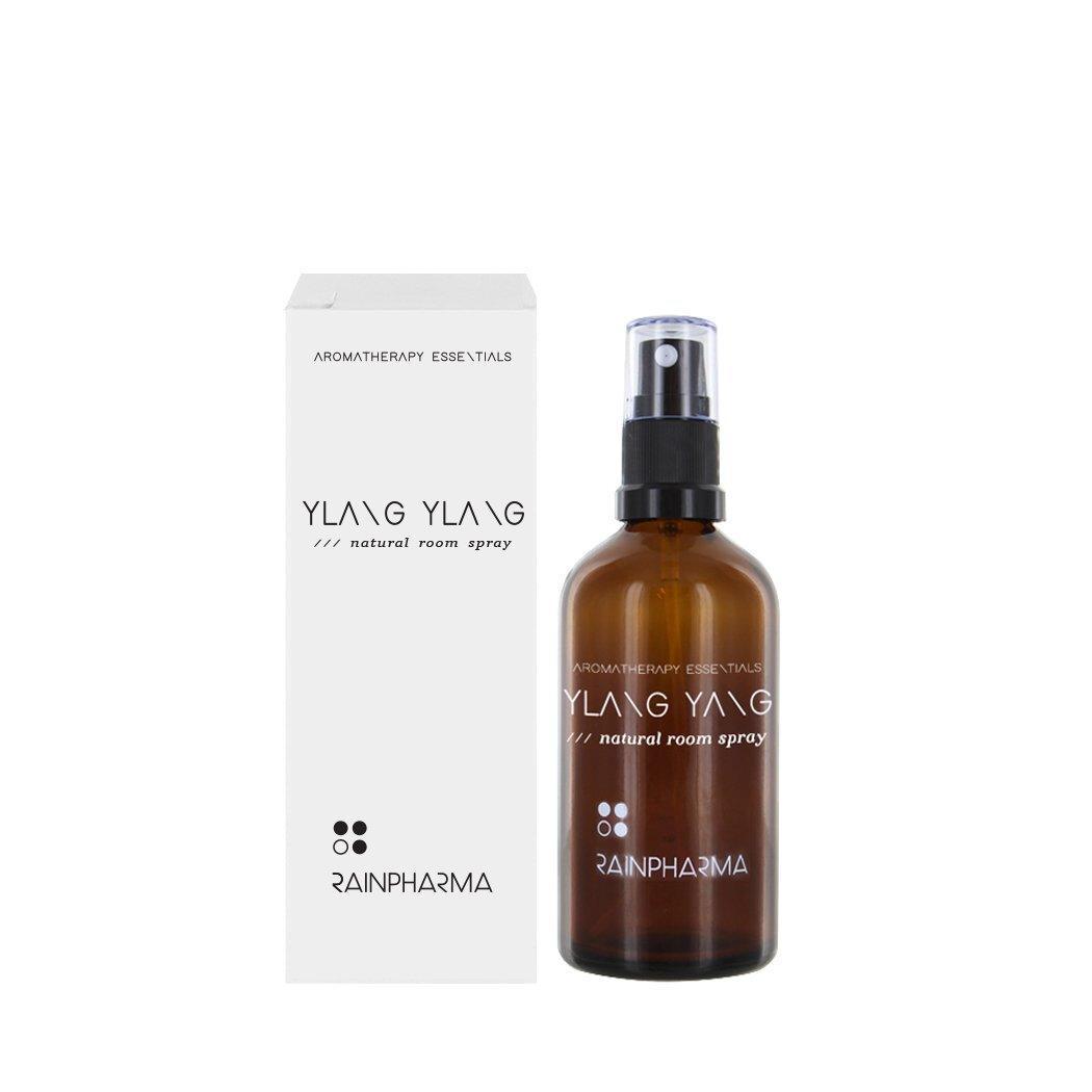 Naast de witte verpakking staat een bruine sprayflacon met de tekst "Natural Room Spray - Ylang Ylang" van RainPharma - een volledig natuurlijke huisgeur verrijkt met essentiële olie.