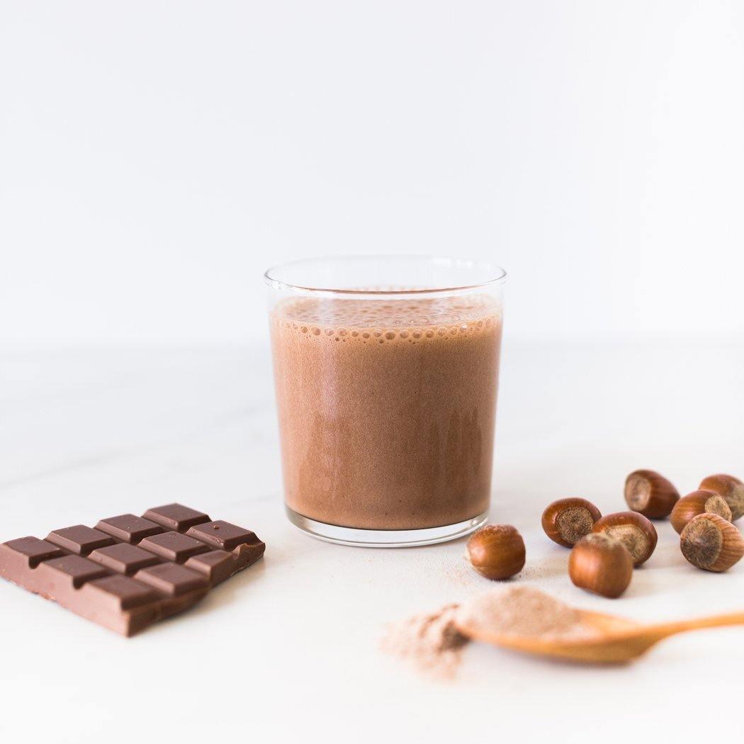 Een glas Noten over Choco Shake op tafel, met een chocolade hazelnoot smaakreep en hazelnoten ernaast, tegen een witte achtergrond.