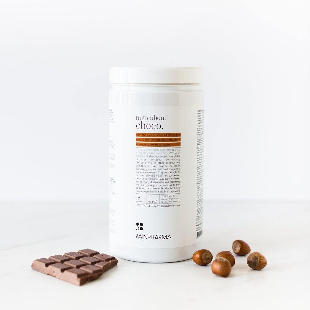 Een container met &quot;Nuts about Choco Shake&quot;, een vegetarisch eiwitrijk Rainpharma-supplement met een reep chocolade-hazelnootsmaak en hazelnoten op een wit oppervlak.
