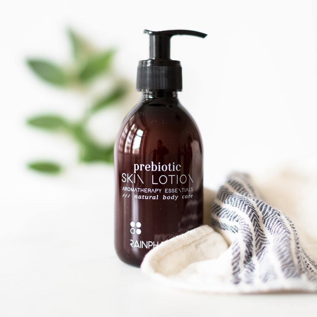 Een donkere RainPharma Prebiotic Skin Lotion fles rust op een wit oppervlak naast een gevouwen doek, met groene bladeren zacht vervaagd op de achtergrond, het vastleggen van de geest van plantaardige huidverzorging.