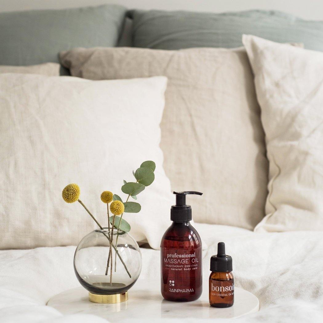 Een ronde glazen vaas met gele bollen en eucalyptus, een pompfles van RainPharma Professionele Massageolie en een druppelflesje staan op een wit dienblad op een bed met beige kussens.