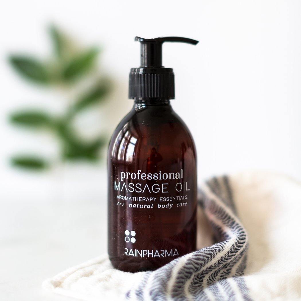 Een bruin RainPharma Massage Oil (Professionele Massageolie) flesje rust op een wit doek, dat aromatherapie oproept, met een vage groene plant op de achtergrond.