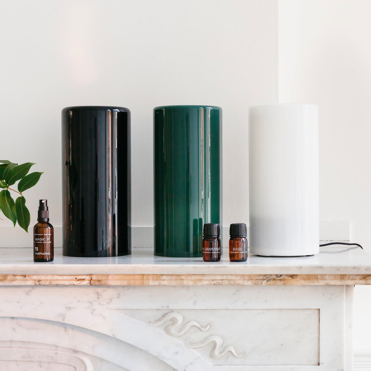 Drie cilindrische luchtreinigers in zwart, groen en wit worden elegant tentoongesteld op een marmeren oppervlak naast drie bruine etherische olieflessen. Het tafereel wordt versterkt door de RainPharma Aroma Diffuser 1,3 L - Aurora Glass Sleeve (kies je kleur), met een elegant glazen koker-ontwerp voor superieure geurverspreiding.