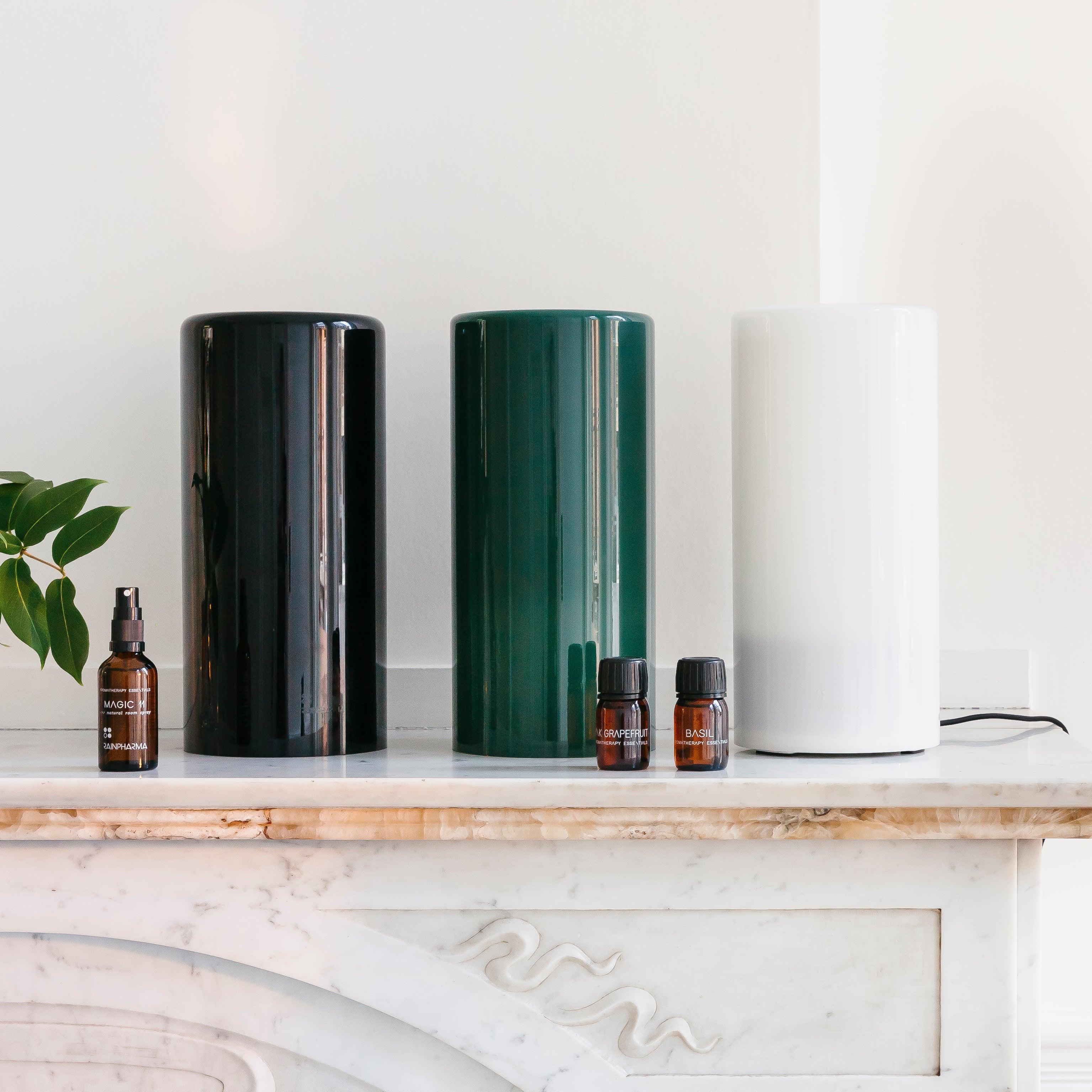 Drie cilindrische luchtreinigers in zwart, groen en wit worden elegant tentoongesteld op een marmeren oppervlak naast drie bruine etherische olieflessen. Het tafereel wordt versterkt door de RainPharma Aroma Diffuser 1,3 L - Aurora Glass Sleeve (kies je kleur), met een elegant glazen koker-ontwerp voor superieure geurverspreiding.