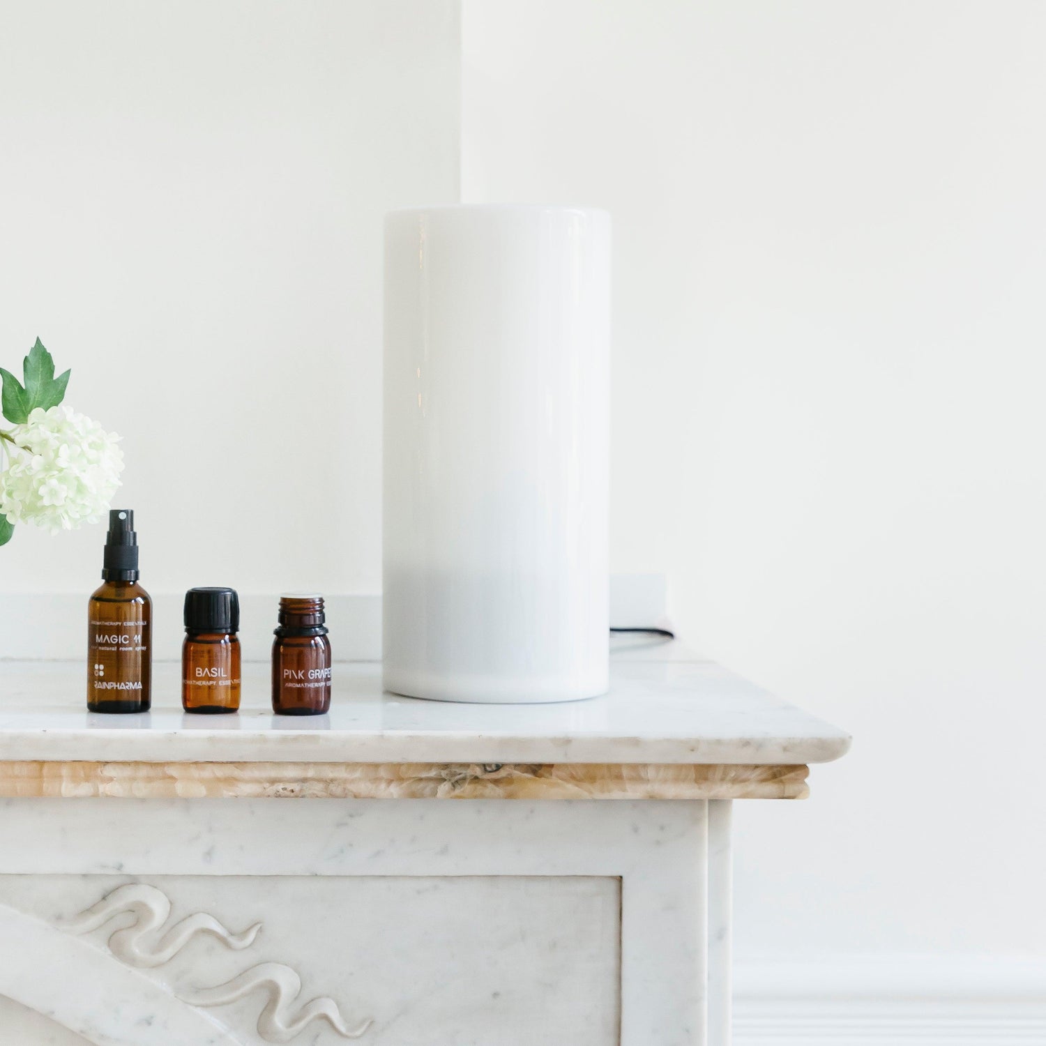 Elegante flesjes etherische olie staan op een marmeren plank naast een witte vaas en bloemen. De geurverspreiding wordt versterkt met de RainPharma Aroma Diffuser 1,3 L - Aurora Glass Sleeve in de buurt.