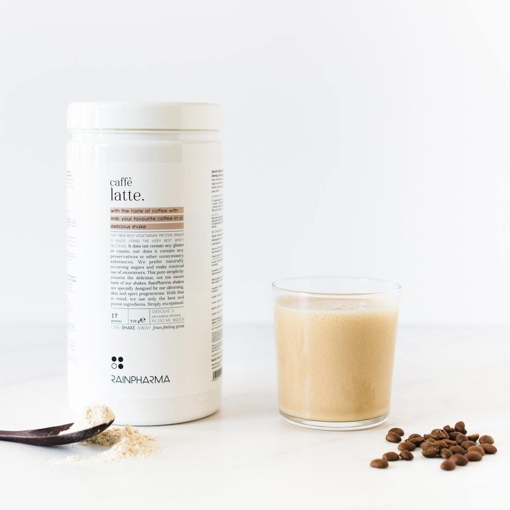 Een bakje met het opschrift &quot;RainPharma Café Latte Shake&quot; naast een glas latte en verspreide koffiebonen op een wit oppervlak, ideaal om te combineren met RainPharma-shakes.