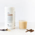 Een bakje met het opschrift "RainPharma Café Latte Shake" naast een glas latte en verspreide koffiebonen op een wit oppervlak, ideaal om te combineren met RainPharma-shakes.