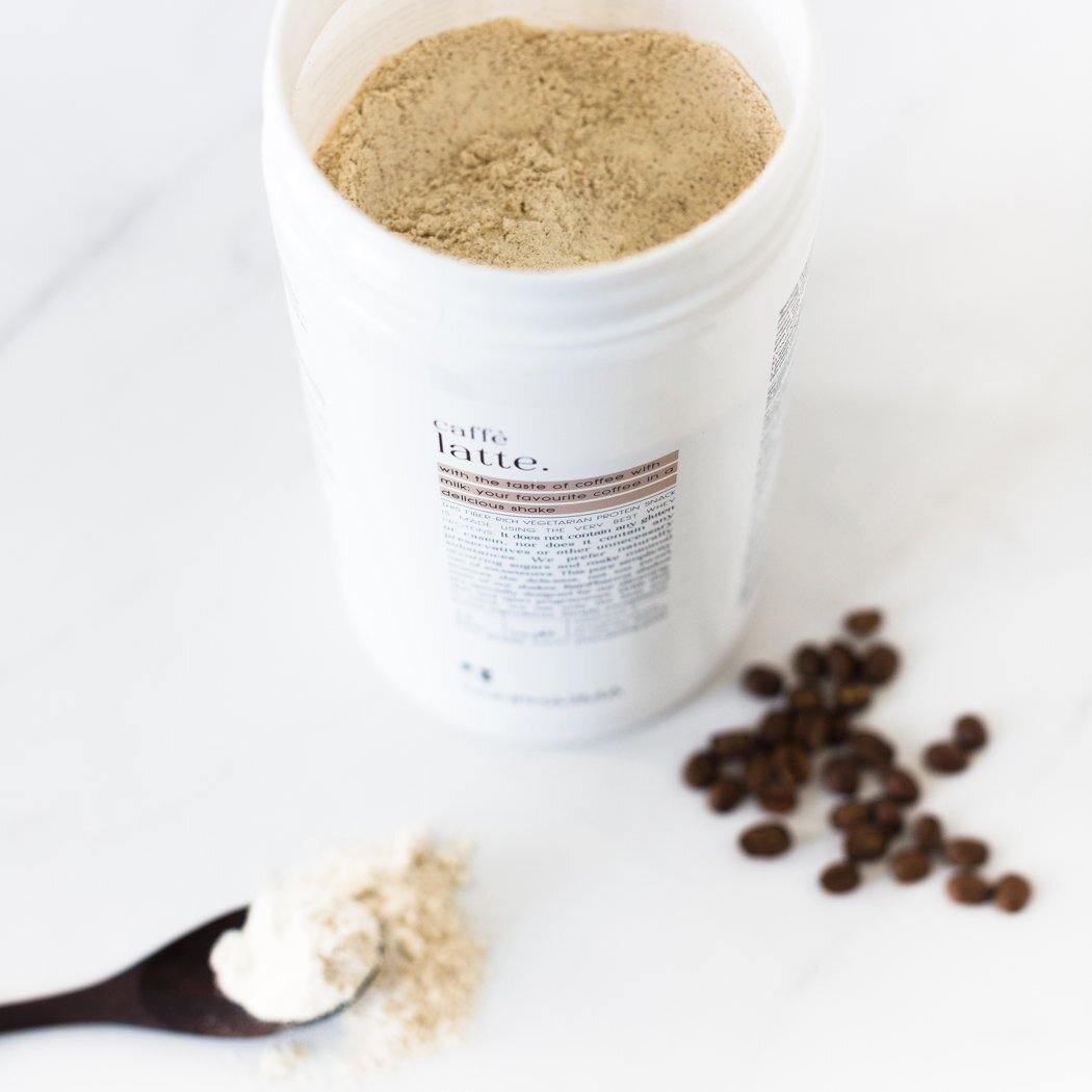 Open een witte container met Rainpharma Café Latte Shake-poeder op een marmeren oppervlak met verspreide koffiebonen en een lepel poeder in de buurt.