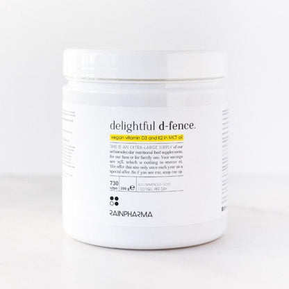 Een witte RainPharma-verpakking met het label &quot;Vitamine D (Delightful D-fence)&quot; bevat een veganistisch vitamine D- en K2-supplement in MCT-olie ter ondersteuning van weerstand, met productinformatie en geel uitgelichte tekst op de voorkant.