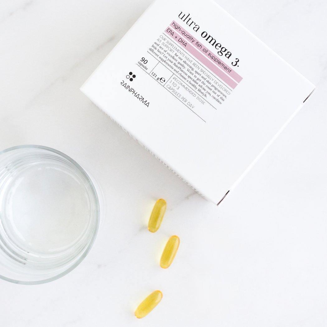 Een witte doos met de tekst "Omega 3 (Ultra Omega 3)" van RainPharma staat naast een leeg glas en drie gele capsules op een wit oppervlak, waarmee de kracht van EPA- en DHA-vetzuren in eersteklas visoliesupplementen wordt benadrukt.