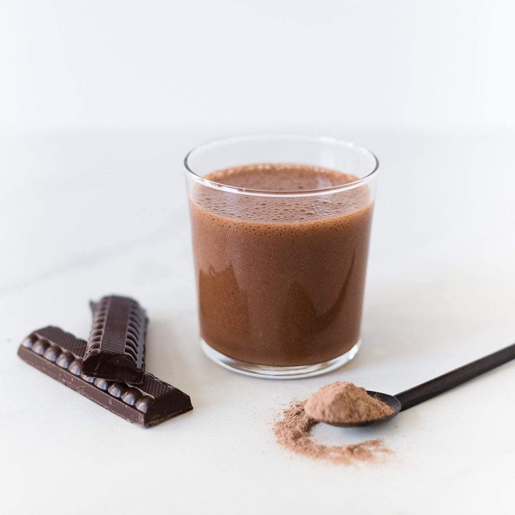 Een glas Rainpharma Raw Chocolate Shake met een reep pure chocolade en een lepel cacaopoeder op een witte ondergrond, zorgen voor een vegetarische eiwitrijke snack.