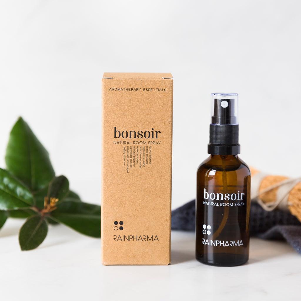 Een bruine doos en een bruine sprayflacon met de tekst "Natural Room Spray - Bonjour" van RainPharma staan op een wit oppervlak met groene bladeren, een schrobber en de frisse sfeer van natuurlijke geuren op de achtergrond.