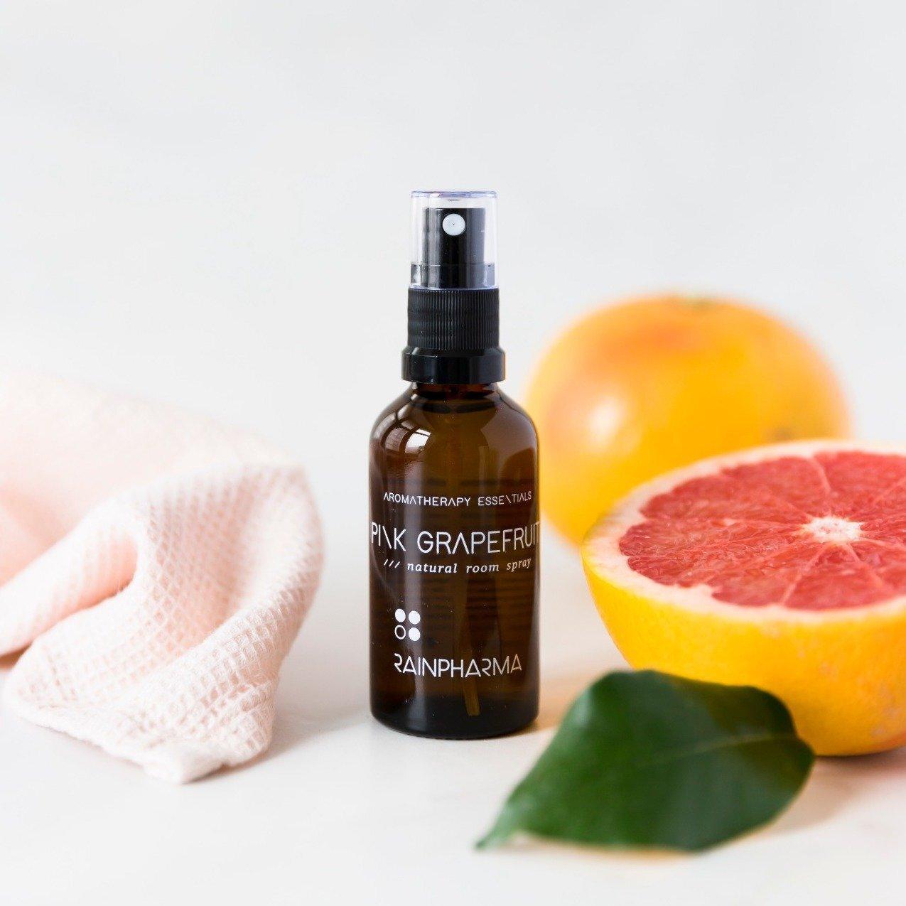 Een fles RainPharma Room Spray Pink Grapefruit staat op een wit oppervlak naast een roze grapefruit, een halve grapefruit, een groen blad en een doek met structuur.