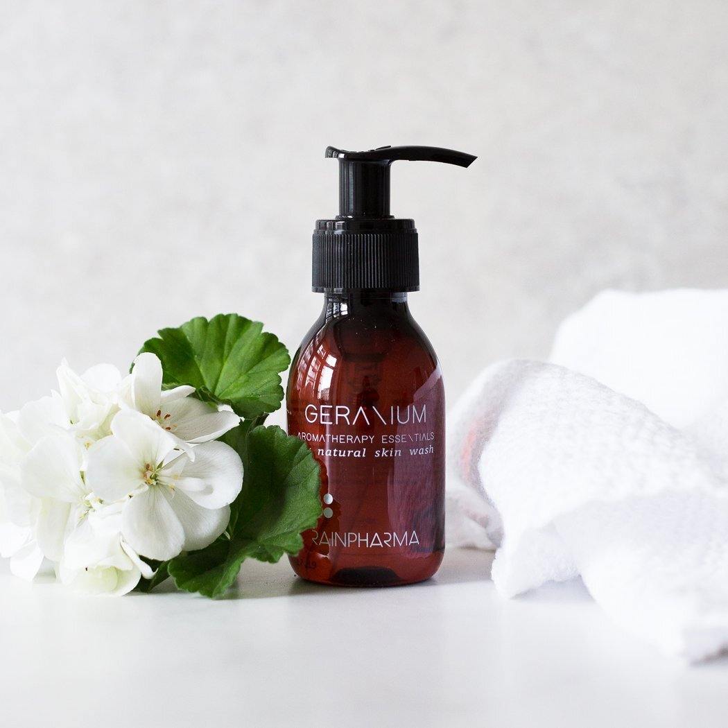 Een fles RainPharma Skin Wash Geranium staat naast witte bloemen en een witte handdoek met textuur op een lichte achtergrond.