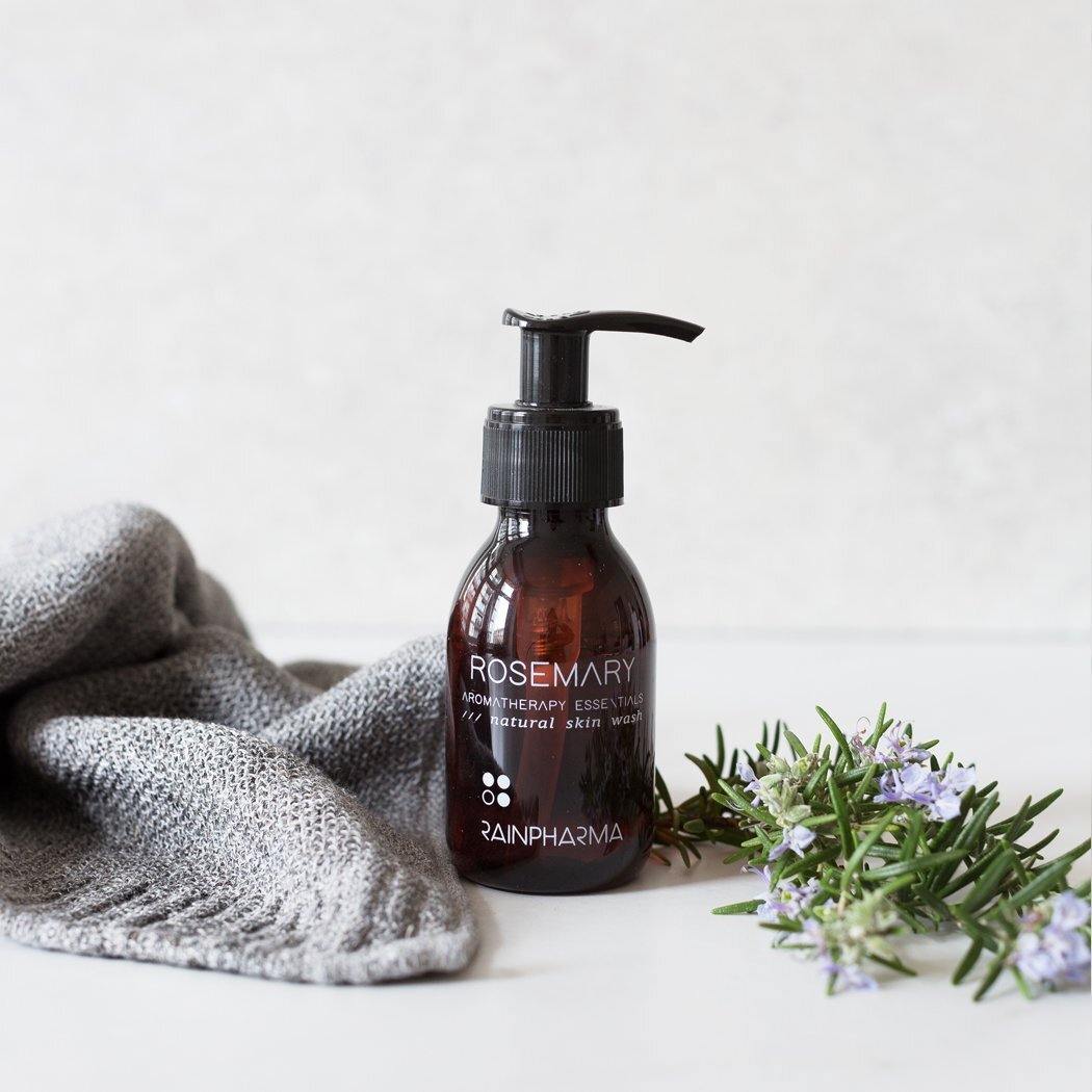 Een bruine pompflacon van RainPharma Skin Wash Rosemary staat naast een grijze gebreide doek en rozemarijntakjes met paarse bloemen op een wit oppervlak, om deze douchegel op plantaardige basis te laten zien.