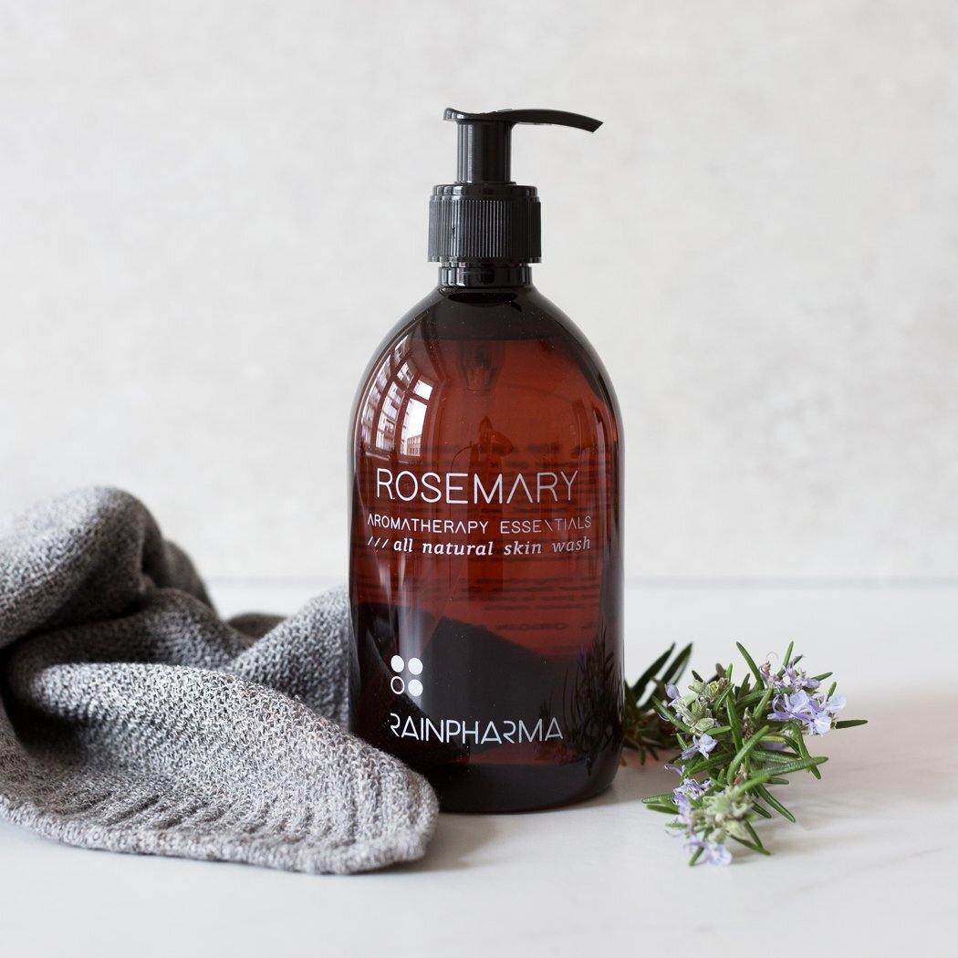 Een bruine pompflacon met RainPharma Skin Wash Rosemary, een douchegel op plantaardige basis, staat naast een grijze handdoek en een takje rozemarijn op een wit oppervlak.