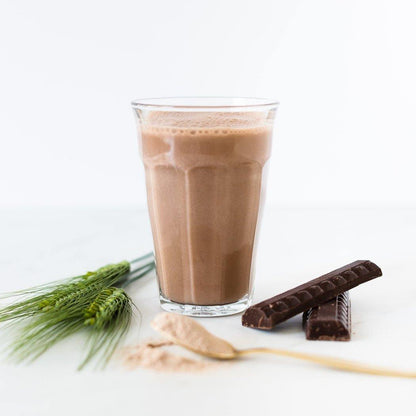 Een glas Rainpharma vaste chocolademilkshake met schuim erop, vergezeld van stukjes chocolade, een lepel en groene tarwestengels op een witte ondergrond.