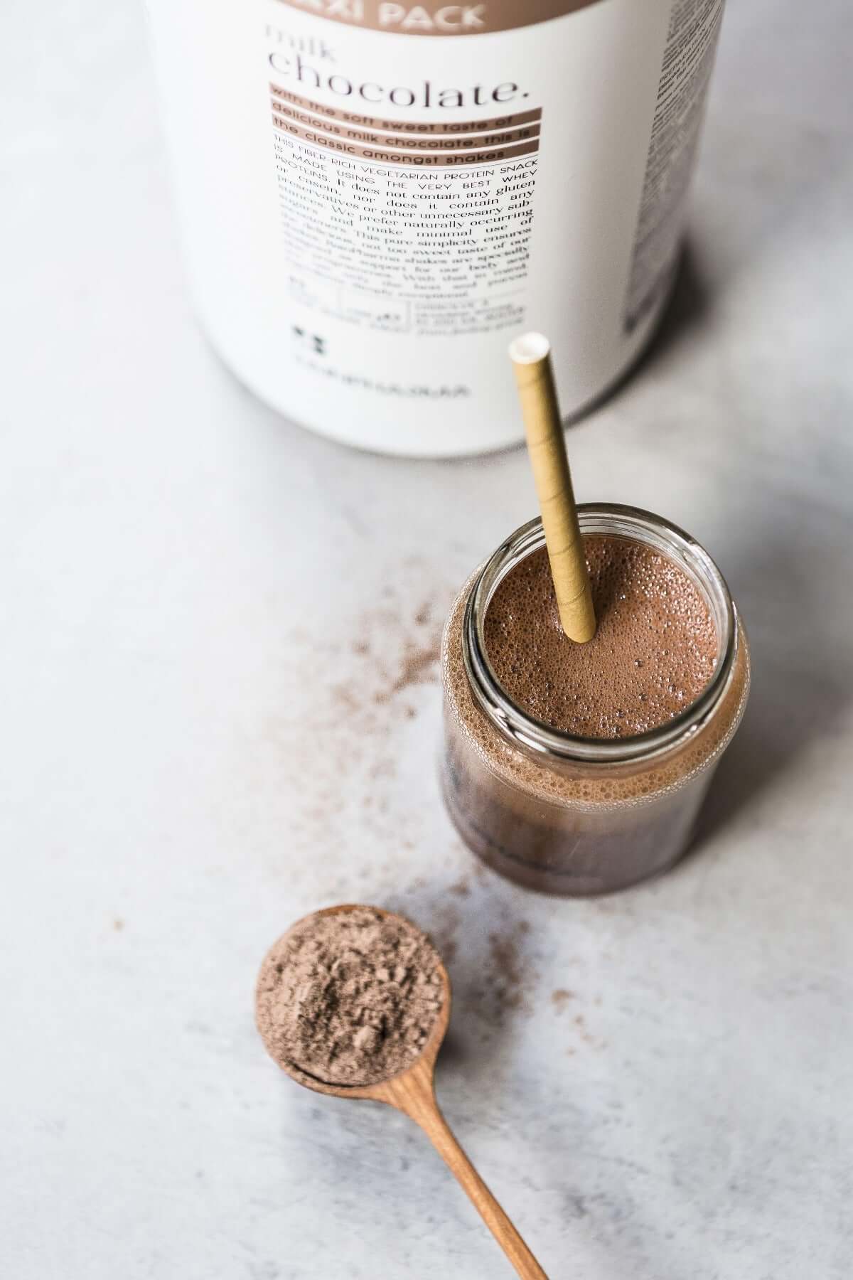 Een potje chocoladeshake met rietje staat naast een houten lepel poeder en een bakje met het opschrift &quot;chocolade&quot;. Deze heerlijke Rainpharma Shake Melkchocolade XL, ideaal als eiwitrijk tussendoortje, is perfect voor wie meer wil.