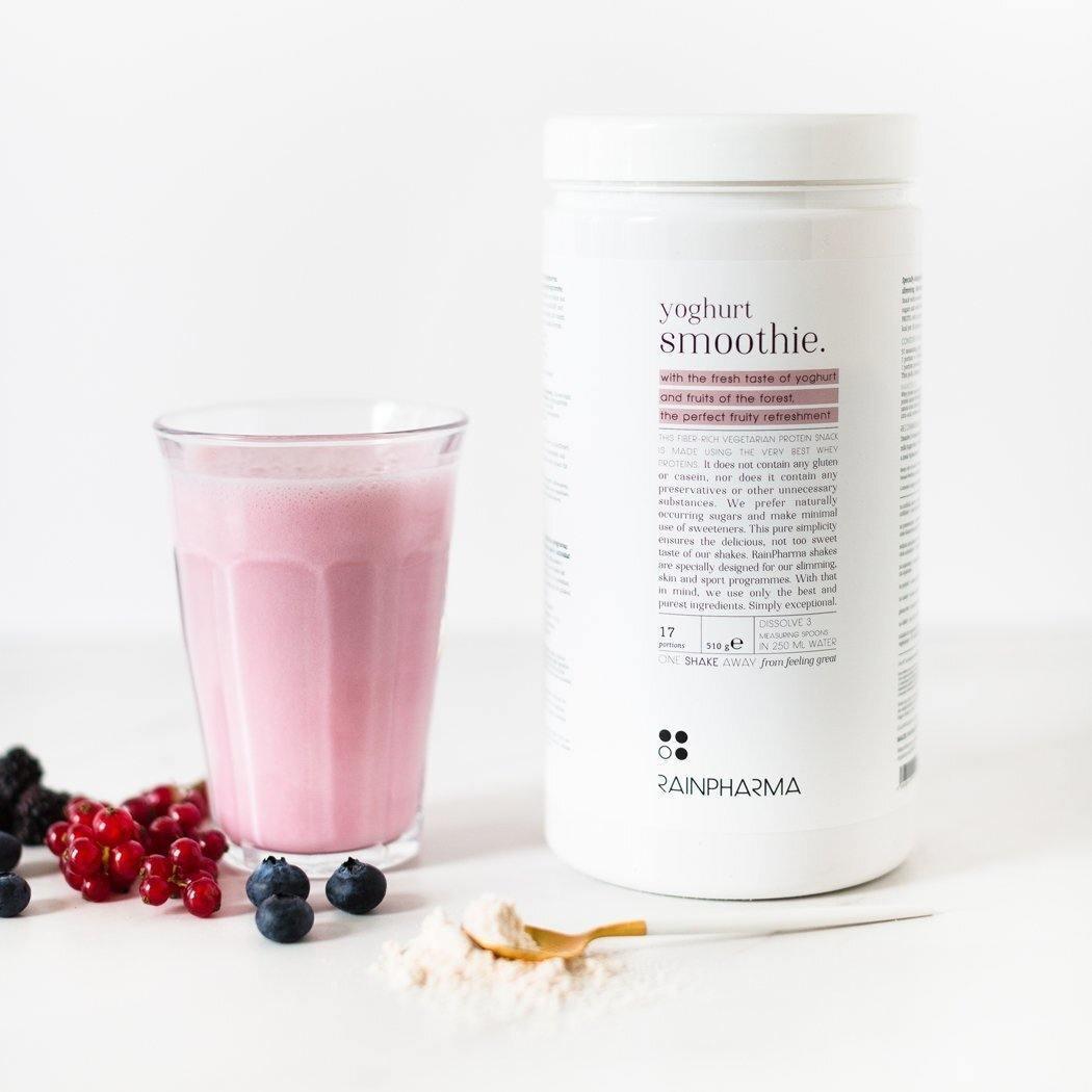 Een glas roze smoothie naast een bakje met het opschrift &quot;RainPharma Shake Yoghurt Smoothie&quot; van Rainpharma, met verspreide bessen en een lepeltje met poeder op een wit oppervlak, zorgt voor een ideaal eiwitrijk tussendoortje.