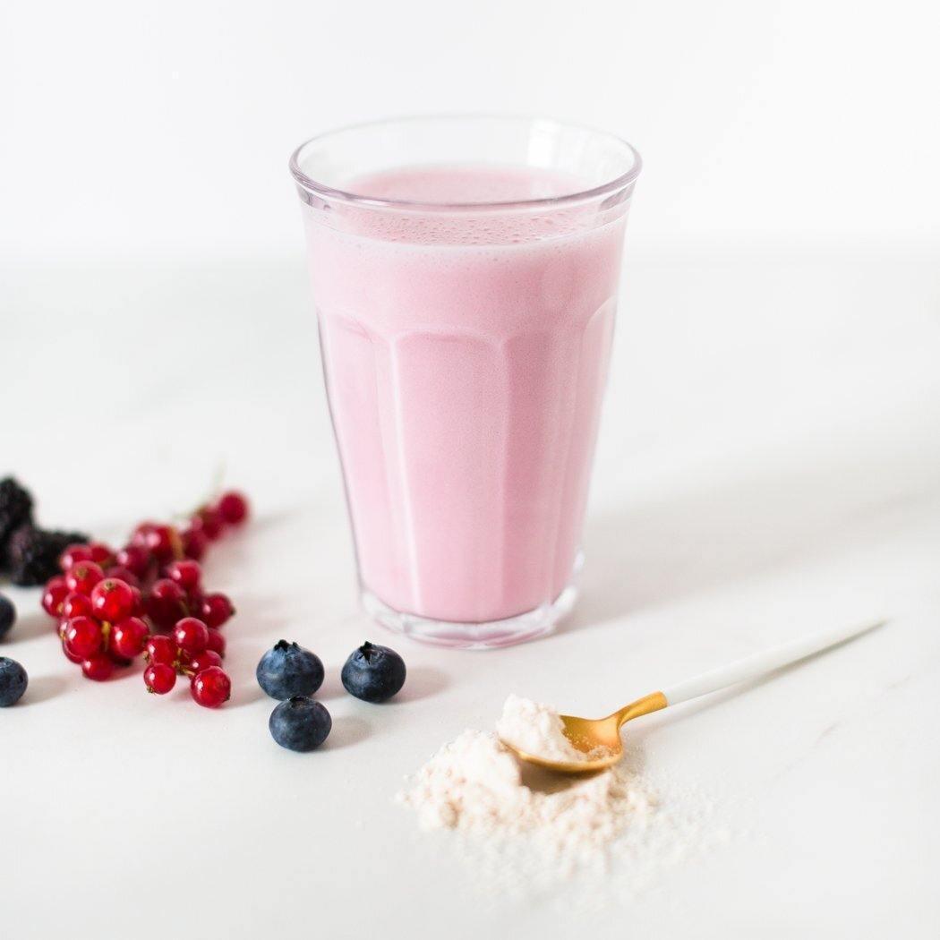 Een glas RainPharma Shake Yoghurt Smoothie, met zijn levendige roze kleur, staat elegant op een wit oppervlak naast een assortiment bessen en een lepel gevuld met poeder.