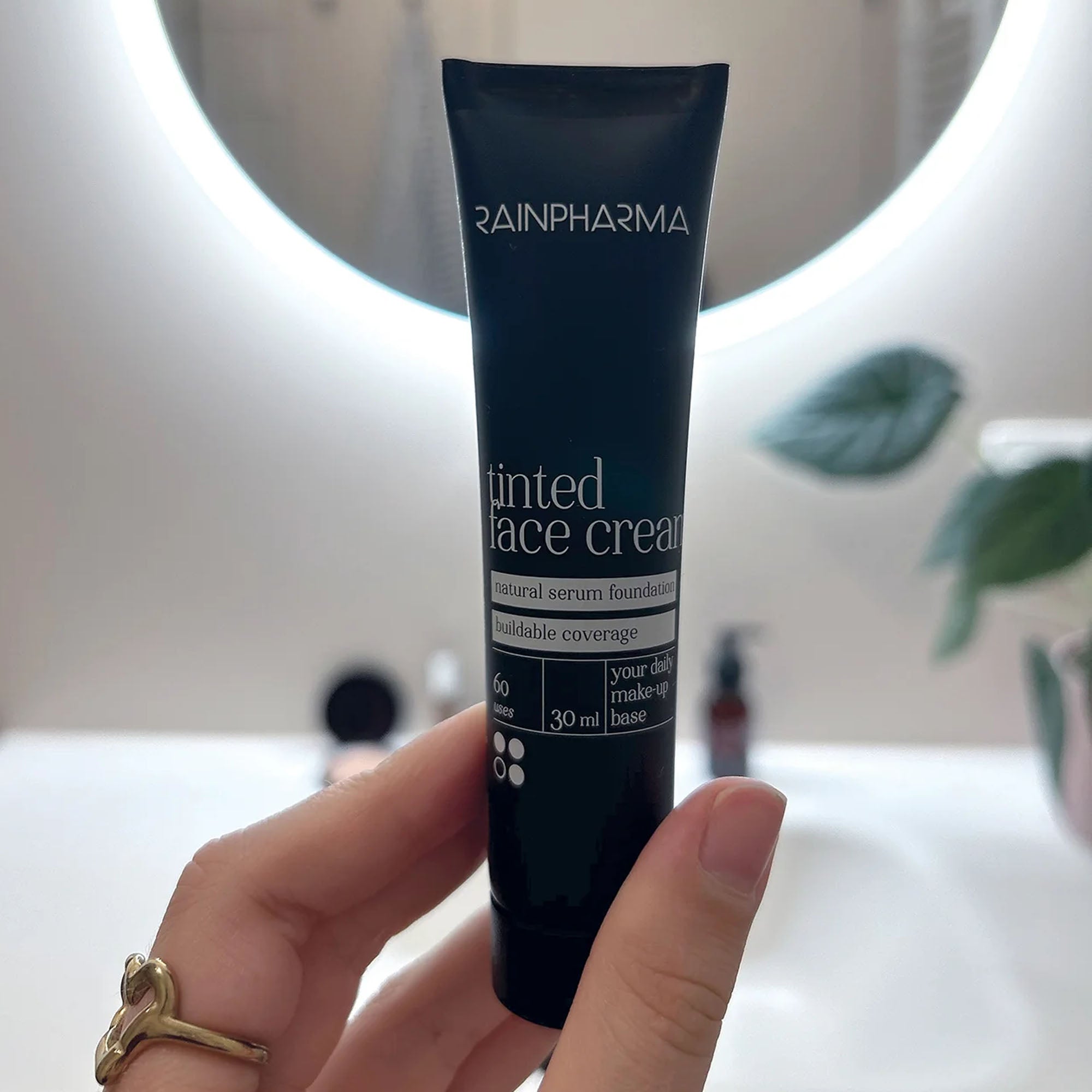 Een hand houdt een tube RainPharma Make Up – Tinted Face Cream voor een ronde spiegel, terwijl vaag groen de achtergrond zachtjes omlijst.