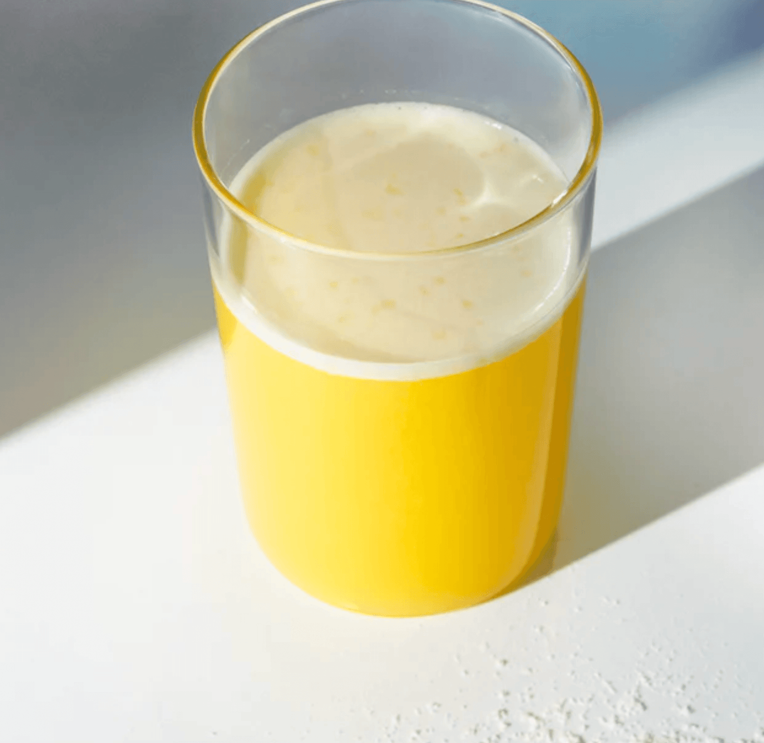 Een glas GOODstuff van Elodie x Rainpharma sinaasappelsap met een schuimige bovenkant, geplaatst op een zonverlicht oppervlak dat zachte schaduwen werpt, verrijkt met collageen als voedingssupplement.