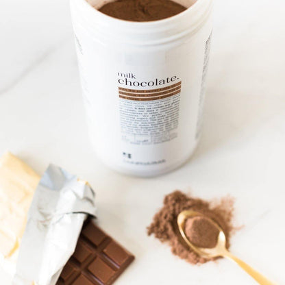 Een grote container Rainpharma Milk Chocolate Shake-poeder naast een onverpakte chocoladereep en een lepel cacao op een witte ondergrond.