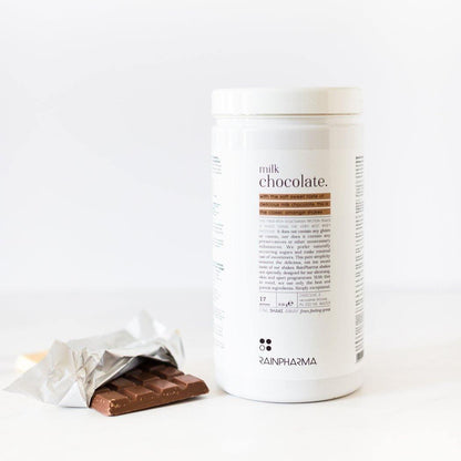 Een witte verpakking met het opschrift &quot;Rainpharma Milk Chocolate Shake&quot; met een etiket met voedingswaarde, naast een gedeeltelijk onverpakte chocoladereep op een wit oppervlak.
