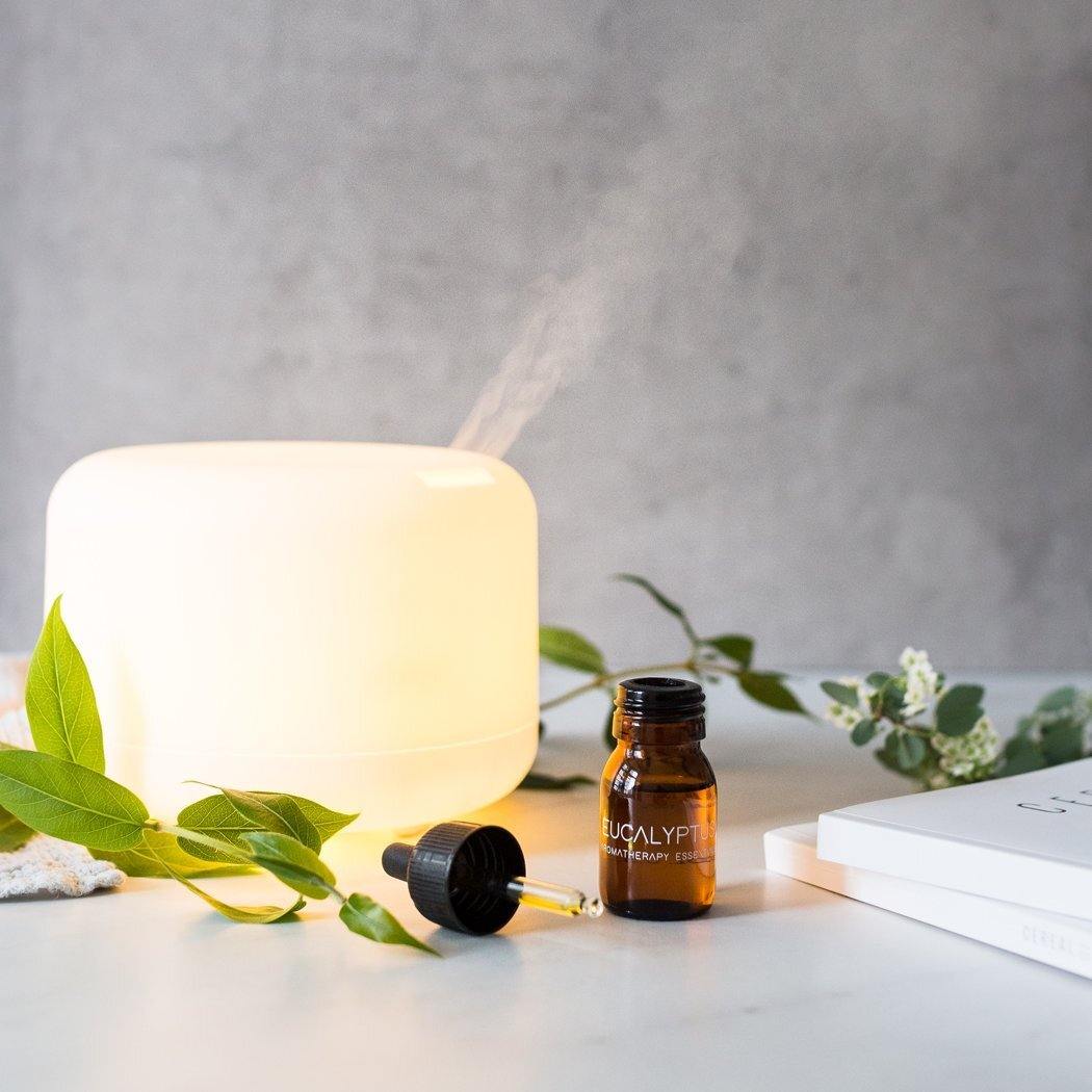 Een RainPharma Aroma Diffuser in het wit verspreidt ultrasone nevel naast een bruine "Eucalyptus"-fles, groene bladeren en een gesloten boek op een licht oppervlak.