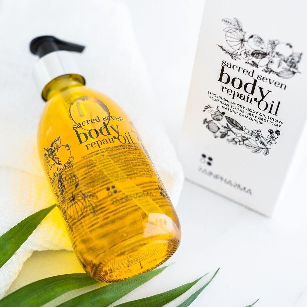 Een pompflacon van RainPharmas Sacred Seven Body Repair Oil staat op een handdoek naast de doos, met groene bladeren ernaast, wat de natuurlijke oorsprong en de premium kwaliteit als lichaamsolie benadrukt.