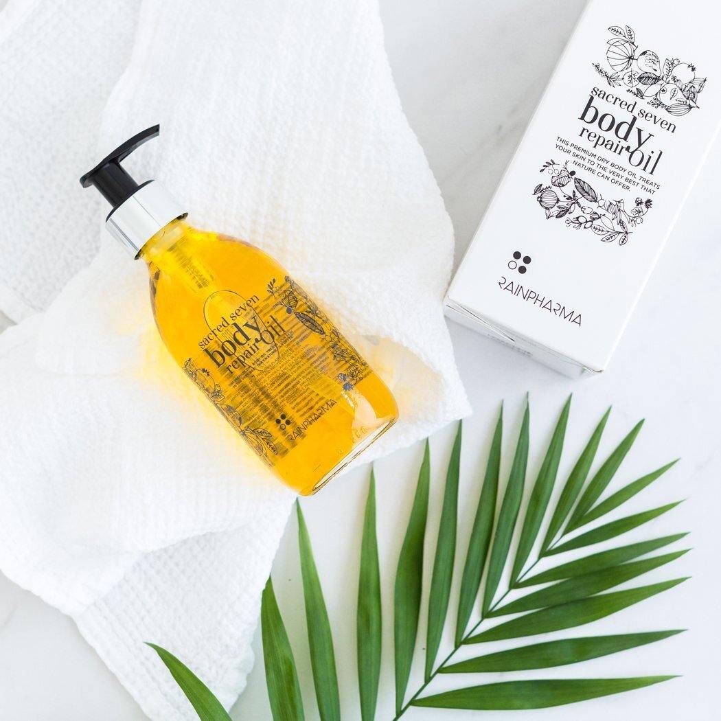 Een pompflacon van RainPharmas Sacred Seven Body Repair Oil, gemaakt met plantaardige ingrediënten, staat op een witte handdoek naast de doos. Een groen palmblad rust elegant op het witte oppervlak, wat de natuurlijke oorsprong benadrukt.