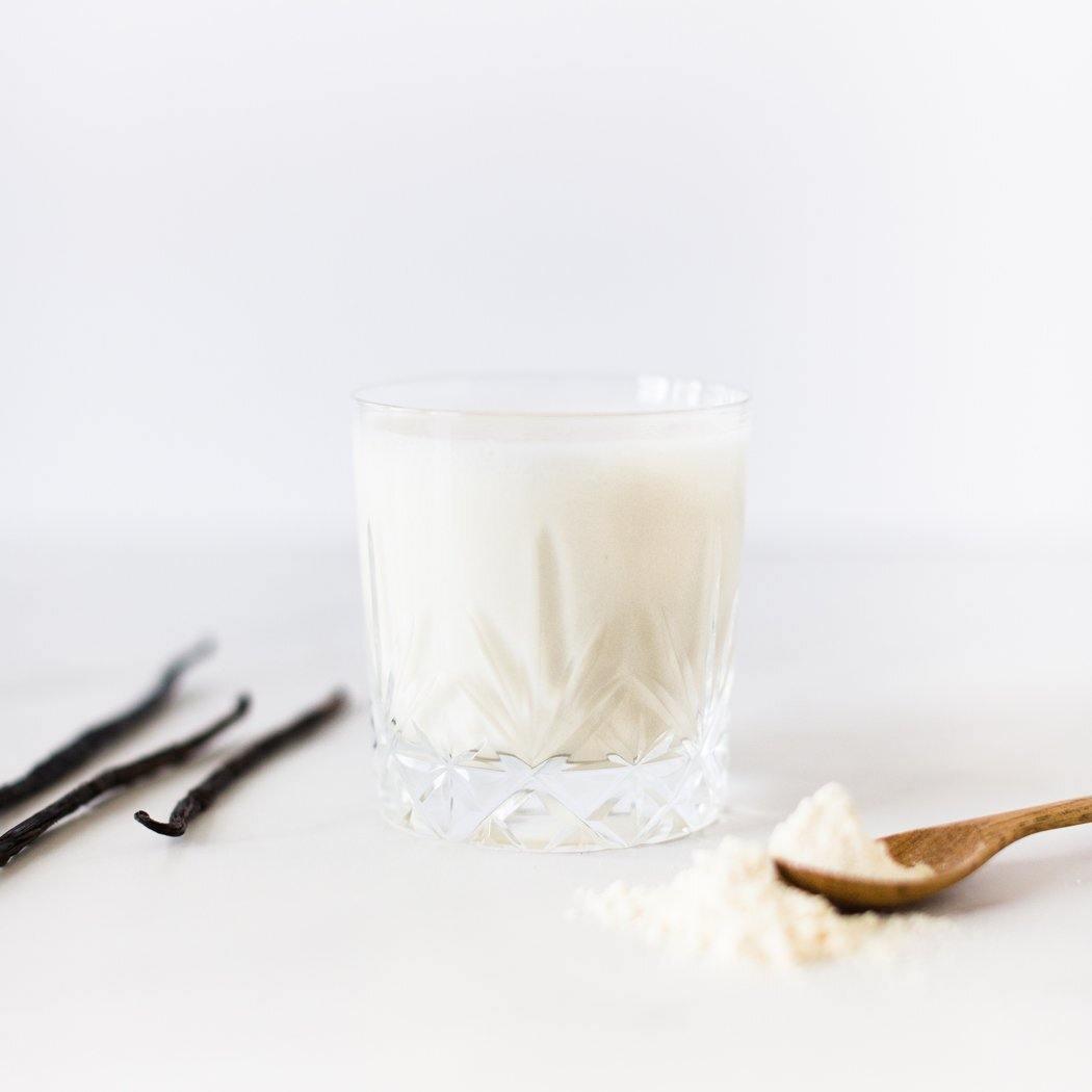 Een glas Simply Vanilla Shake van Rainpharma op een witte achtergrond met vanillestokjes en een lepel suiker in de buurt.