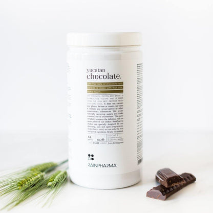 Een Rainpharma Vegan Chocolate Shake bodyscrubcontainer op een witte ondergrond, vergezeld van stukjes chocolade en groene planten.