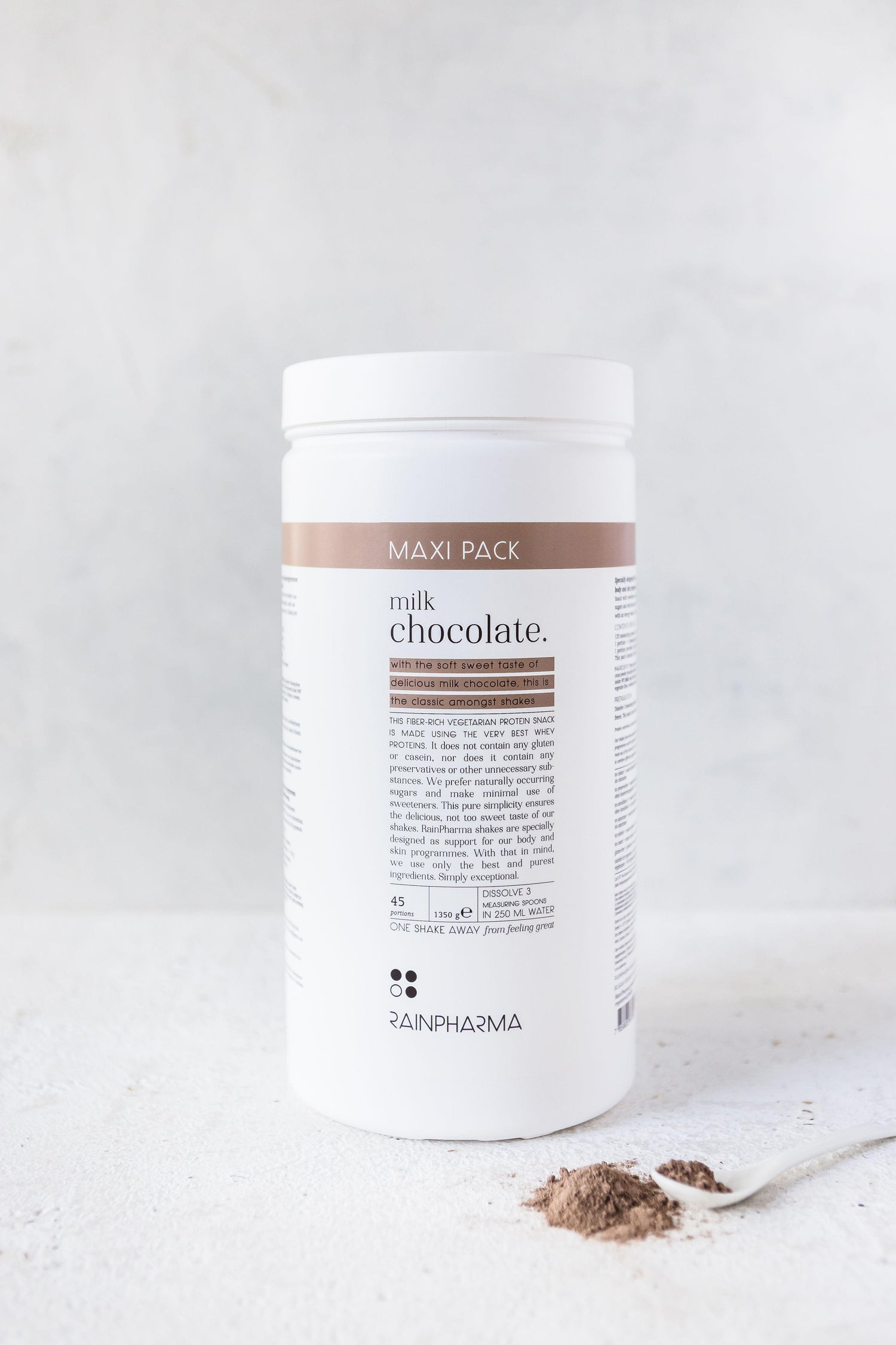 Een grote witte container met het opschrift &quot;XL Shake Milk Chocolate - PRE-ORDER (levering juni 24)&quot; van Rainpharma, met voedingsinformatie, geplaatst op een lichte achtergrond, naast een lepel met chocoladepoeder.