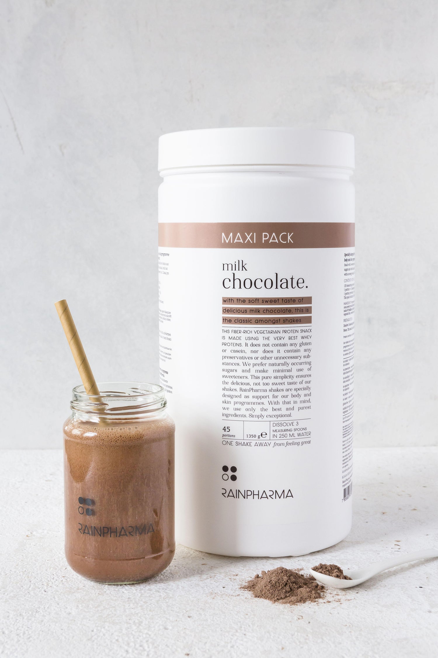 Een grote container XL Shake Milk Chocolate van Rainpharma naast een glas chocoladedrank met rietje, op een lichte achtergrond.