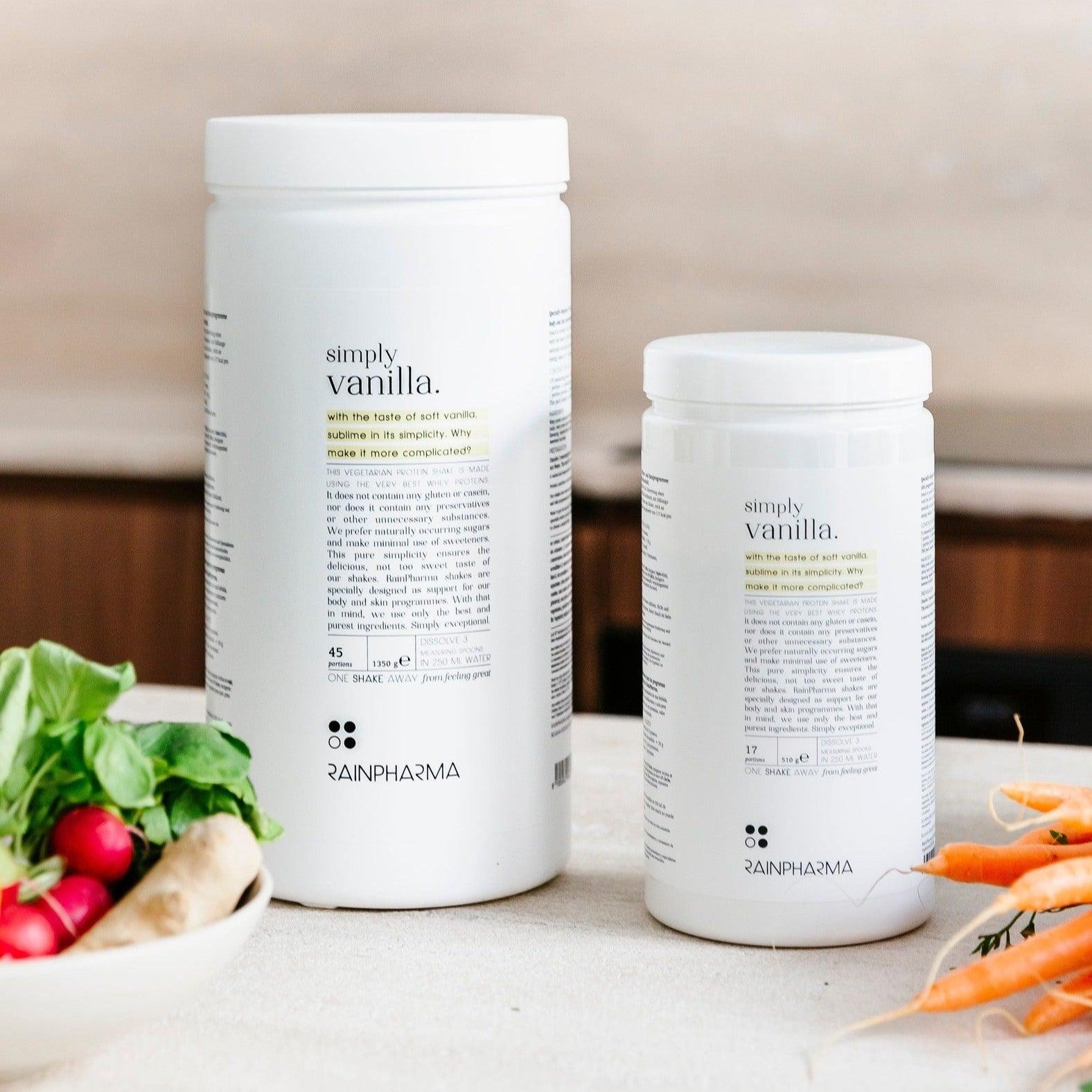 Twee containers Rainpharma XL Shake Simply Vanilla - Limited Edition voedingssupplement op een aanrecht, omringd door verse groenten.